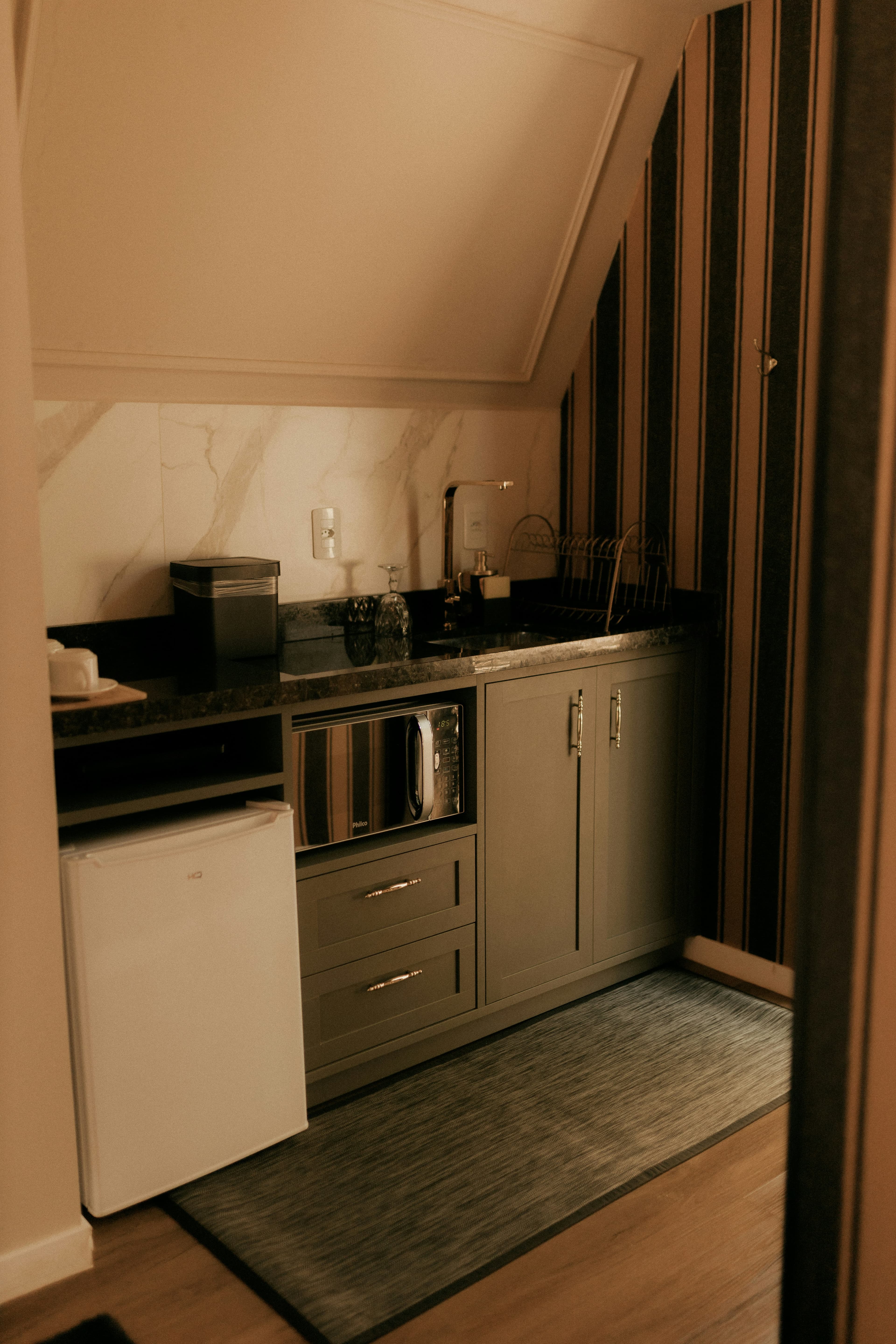 Compact kitchenette with muted cabinetry, marble backsplash, gold fixtures, and stone counter